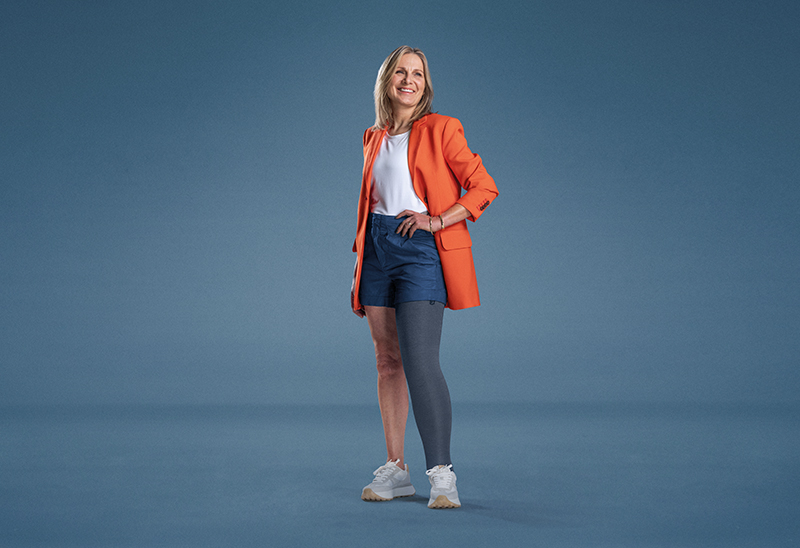 JOBST ambassador Julie is standing on a blue background, dressed in an orange blazer and grey compression garment on her leg. JOBST ambassador Julie is standing on a blue background, dressed in an orange blazer and grey compression garment on her leg.